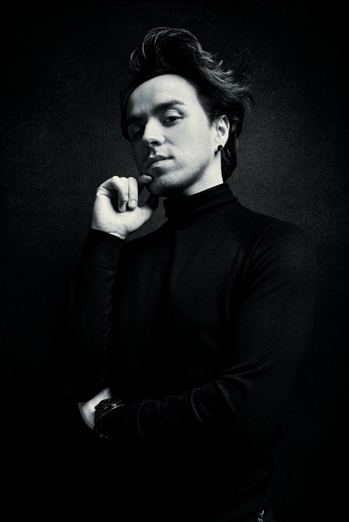 Czarno-biały portret mężczyzny w golfie, z dłonią przy brodzie i zegarkiem na nadgarstku, na ciemnym tle. Stylizowane oświetlenie podkreśla rysy twarzy.