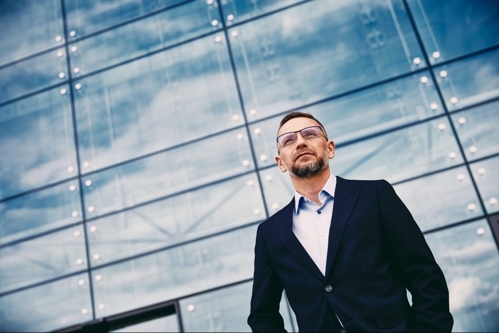Mężczyzna w garniturze i okularach spogląda w górę na szklaną fasadę budynku, w której odbija się niebo. Ujęcie z dołu, podkreślające nowoczesną architekturę.