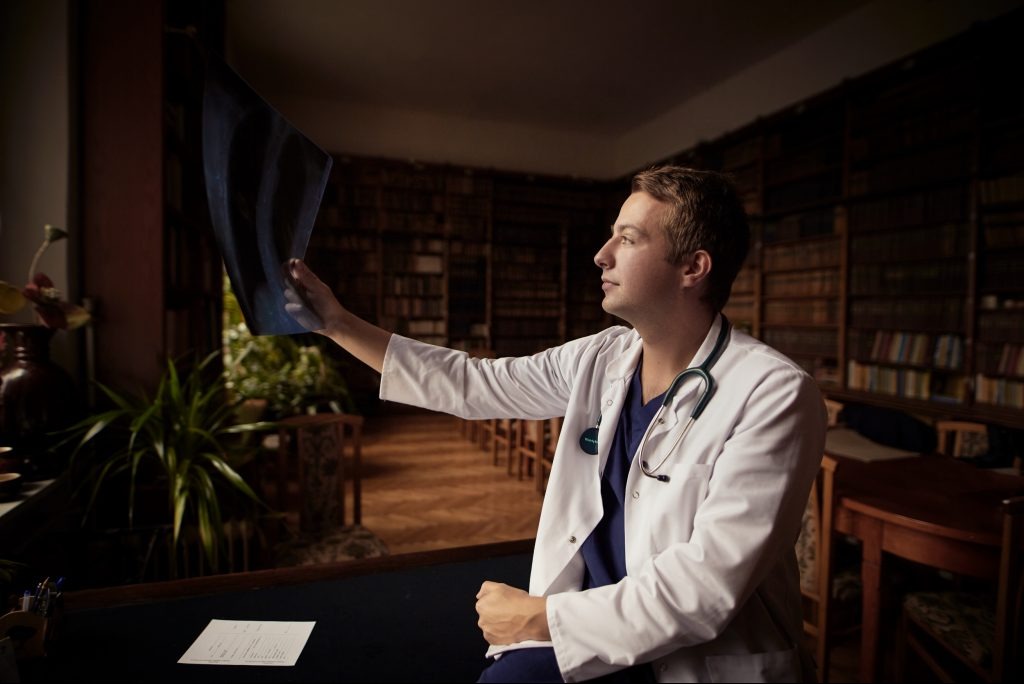 Lekarz w białym kitlu analizuje zdjęcie rentgenowskie w gabinecie z biblioteczką. Stetoskop na szyi, dokumenty na biurku. Delikatne, naturalne oświetlenie.