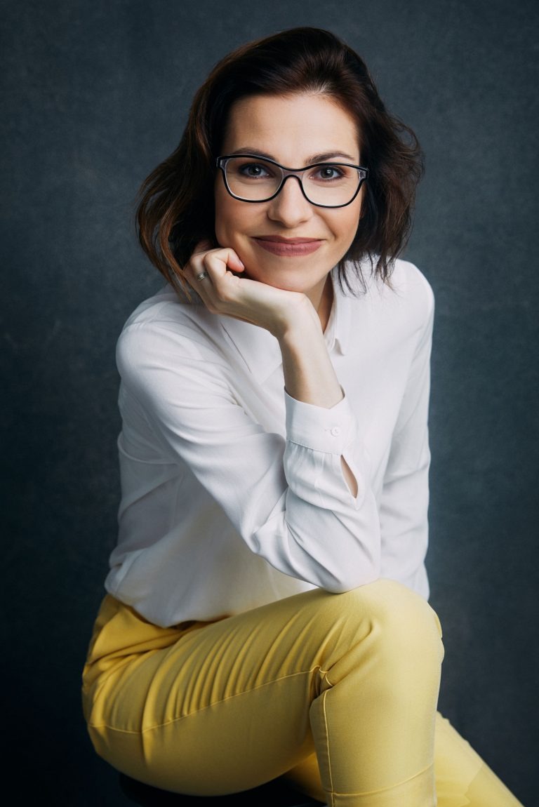 Portret kobiety w okularach, białej koszuli i żółtych spodni na ciemnym tle. Uśmiechnięta, z dłonią pod brodą, patrzy w obiektyw. Profesjonalna sesja.