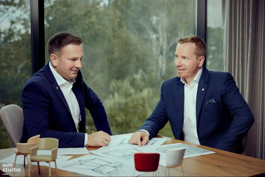 Dwóch mężczyzn w garniturach omawia projekt architektoniczny przy drewnianym stole z modelami krzeseł, na tle okna z widokiem na zieleń.