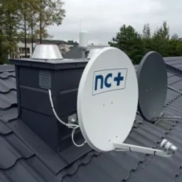 Dwie anteny satelitarne, biała z logo nc+ i szara, zamontowane na ciemnoszarym dachu z blachodach&oacute;wki, obok komina z metalowym daszkiem.