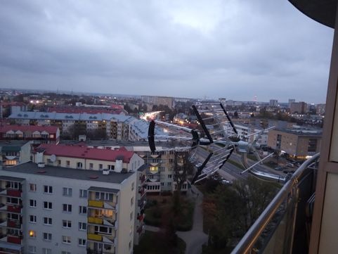 Antena telewizyjna zamontowana na balkonie z widokiem na miasto w pochmurny dzień, widoczne bloki mieszkalne i ulice z perspektywy balkonu.