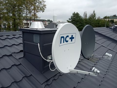 Dwie anteny satelitarne, biała z logo nc+ i szara, zamontowane na ciemnoszarym dachu z blachodachówki, obok komina z metalowym daszkiem.