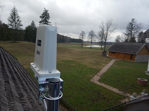 Biała antena z logo firmy Plus zamontowana na dachu z widokiem na wiejski krajobraz z drewnianym budynkiem i stawem w tle przy pochmurnej pogodzie.