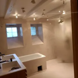 Łazienka w trakcie wykańczania z mikrocementem na ścianach i podłodze, widoczne dwa okna w głębokich wnękach, sufit z drewnianymi belkami i zamontowanymi lampami, zabudowa umywalkowa z drewnianymi...