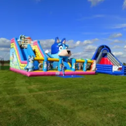 Dwa dmuchane zamki w stylu kreskówki Bluey na trawniku, z błękitnym niebem i chmurami w tle.