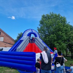 Jacek Cieliński - Dmuchana zjeżdżalnia w kolorach niebieskim, szarym i czerwonym, z dziećmi na górze, ustawiona na trawie obok ceglanego budynku i drzew. Widoczni ludzie obserwujący zabawę.