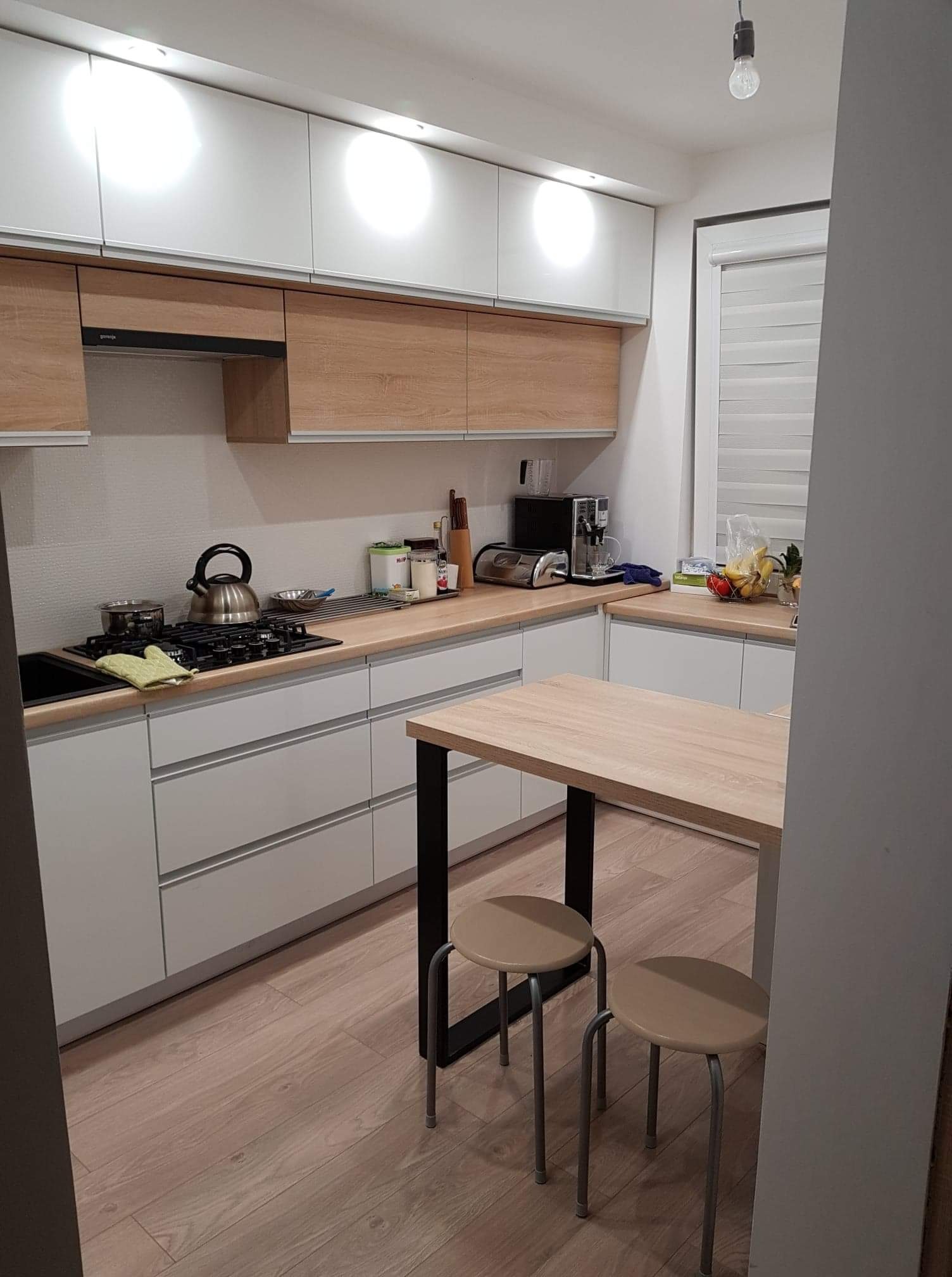 Nowoczesna kuchnia z białymi frontami i drewnianymi akcentami, widoczny stół z hokerami, czajnik na płycie grzewczej i owoce na blacie.