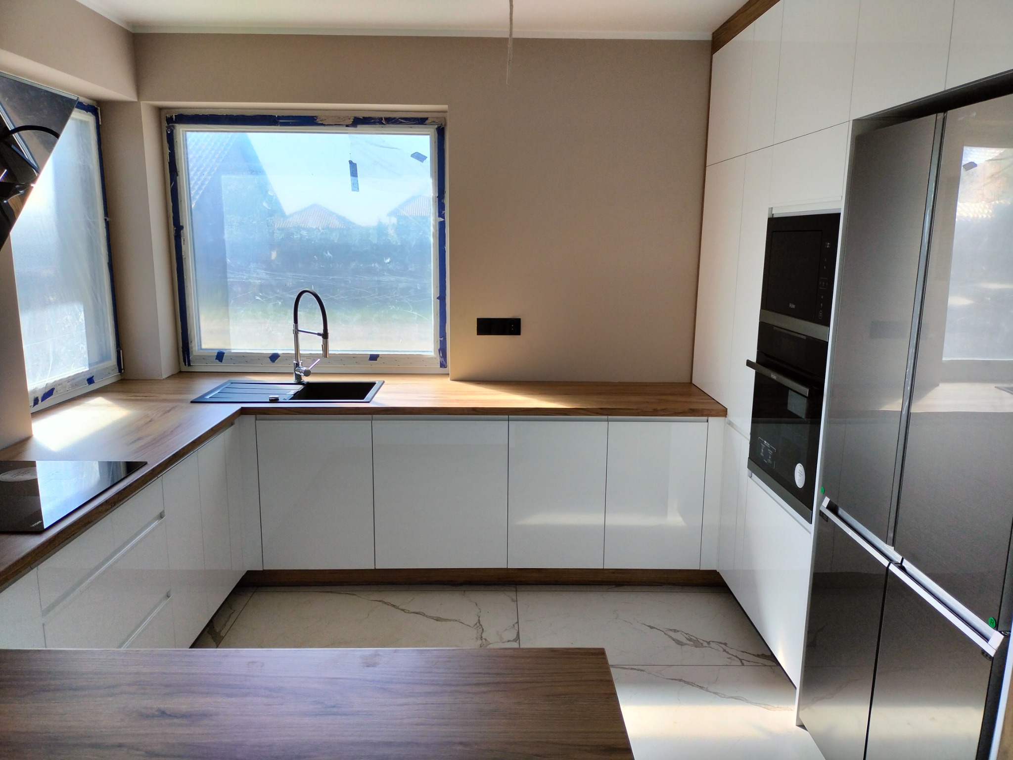 Nowoczesna, jasna kuchnia z białymi szafkami i drewnianym blatem, widok na okno z czarną armaturą i zlewozmywakiem, marmurowe płytki na podłodze, zabudowana lodówka i piekarnik.