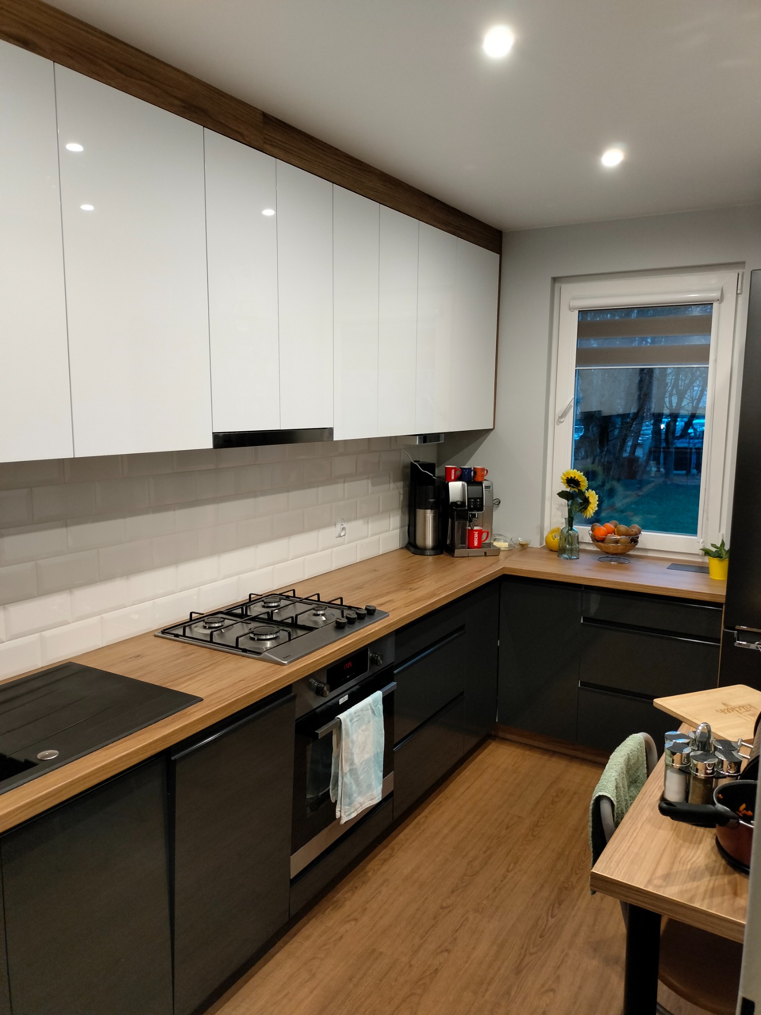 Nowoczesna kuchnia z białymi szafkami górnymi i czarnymi dolnymi, drewniany blat, okno z widokiem na zieleń.