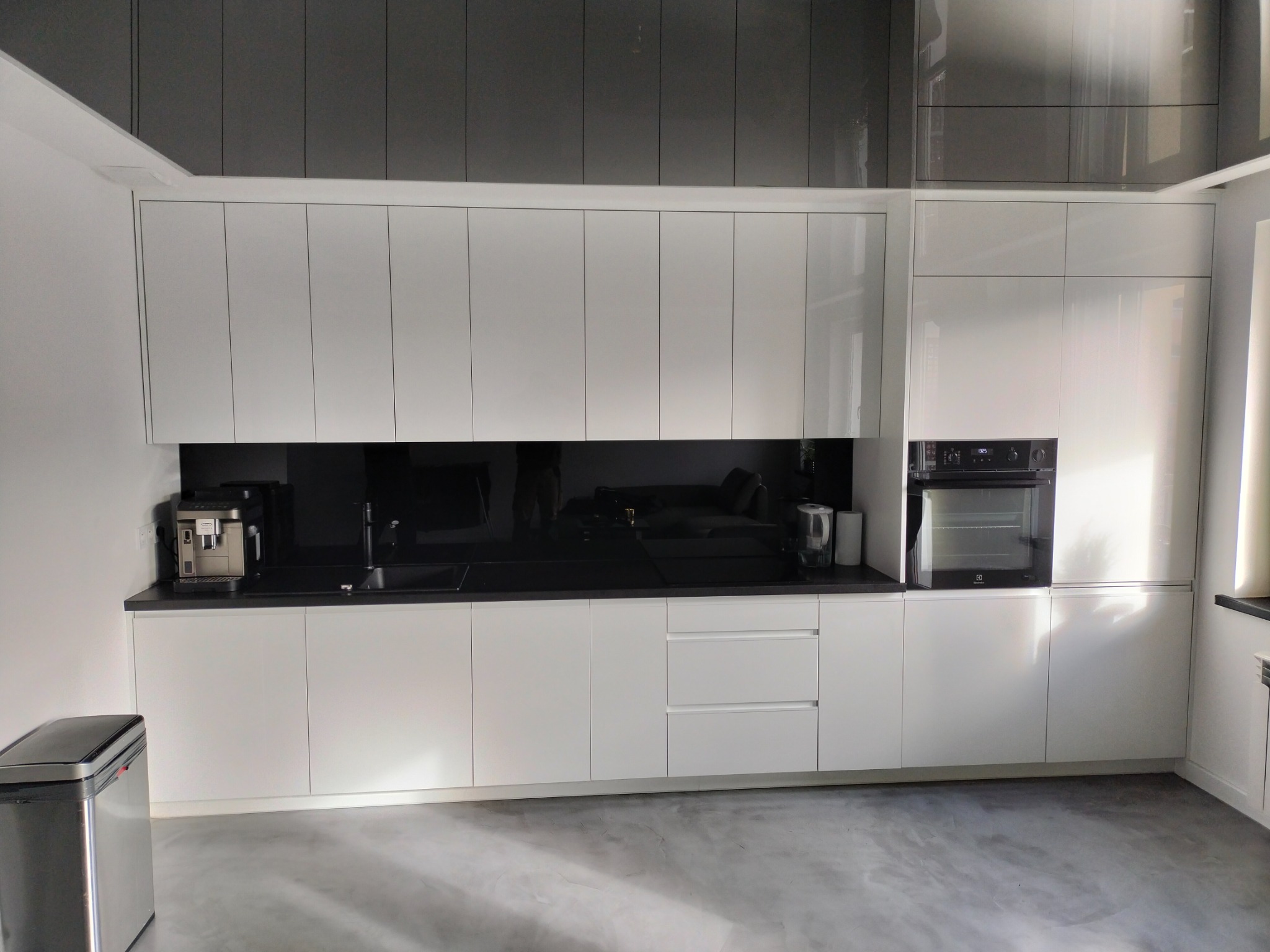 Nowoczesna kuchnia z białymi, lakierowanymi frontami, czarnym blatem i panelem nad blatem, widoczny ekspres do kawy i piekarnik.