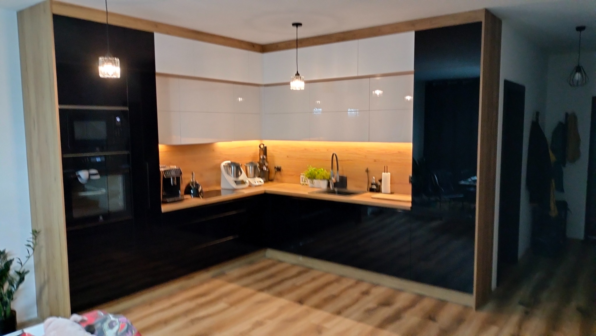 Nowoczesna kuchnia z białymi szafkami górnymi i czarnymi dolnymi, drewniany blat i oświetlenie LED pod szafkami, widoczne sprzęty AGD i lampy wiszące.