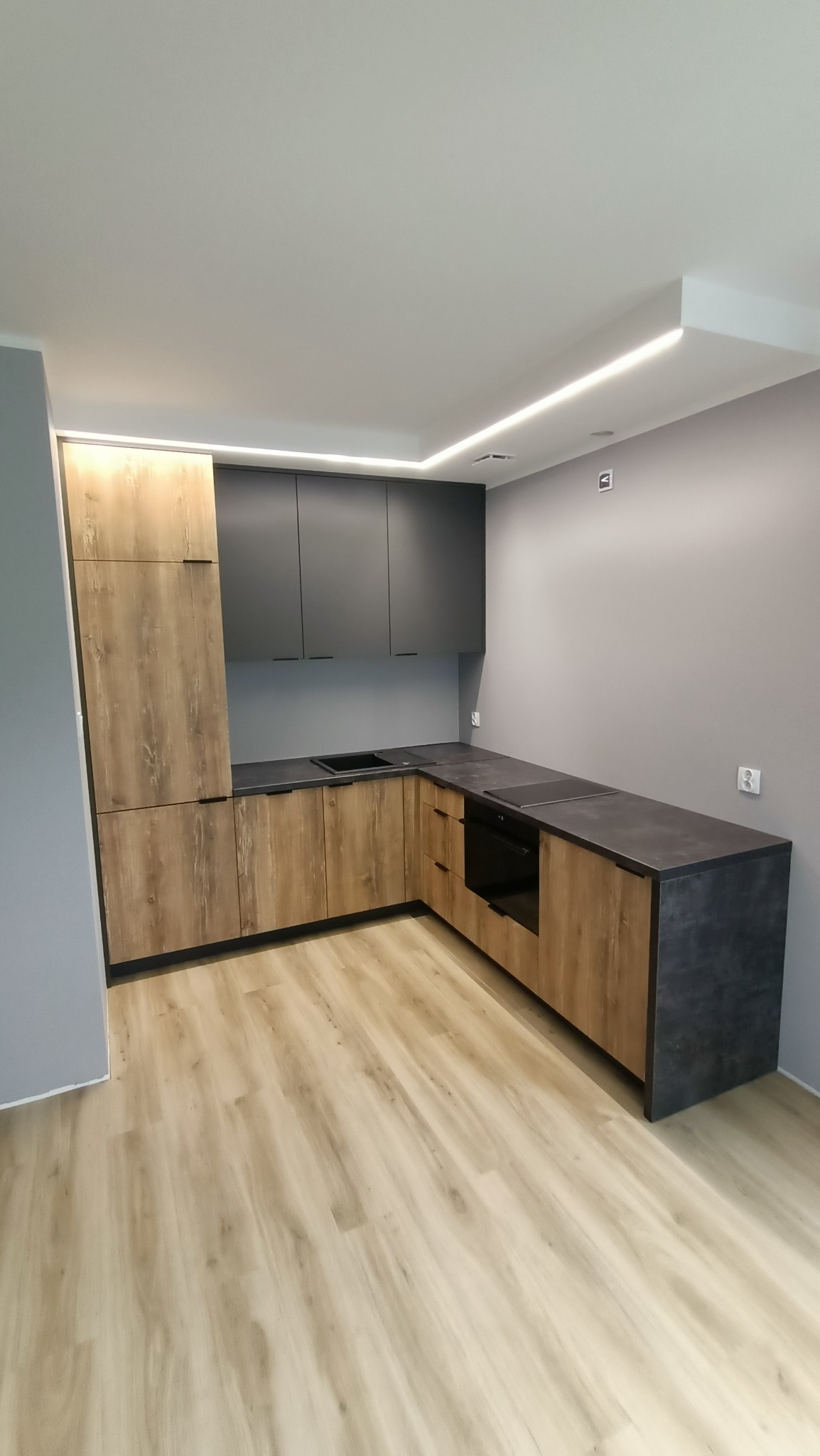 Nowoczesna kuchnia z drewnopodobnymi frontami szafek dolnych i grafitowymi szafkami górnymi, czarny blat, oświetlenie LED podszafkowe i na suficie, widok na narożnik z piekarnikiem.