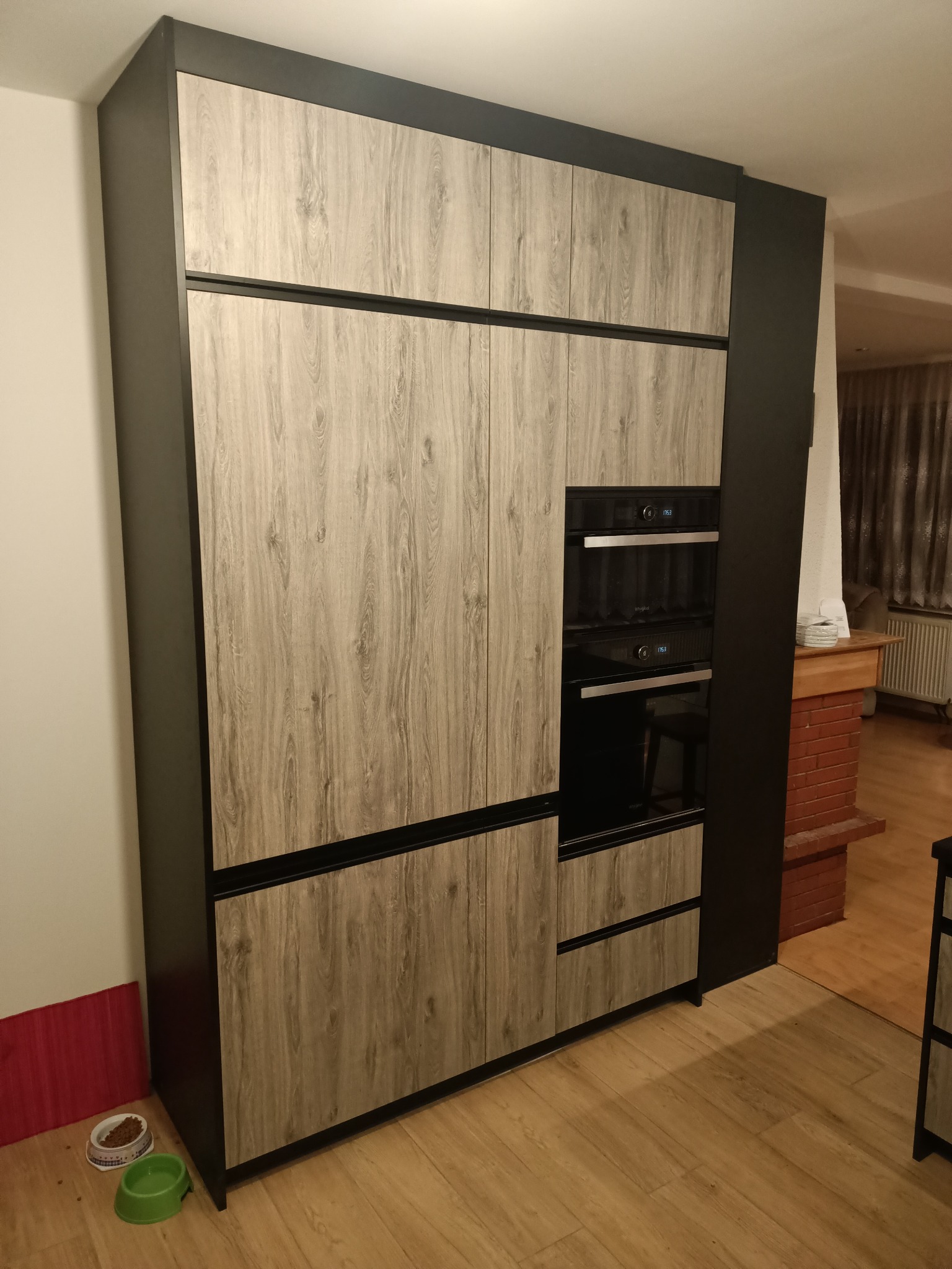 Wysoka zabudowa kuchenna z fornirem imitującym drewno i wbudowanymi piekarnikami, widoczne miski dla zwierząt.