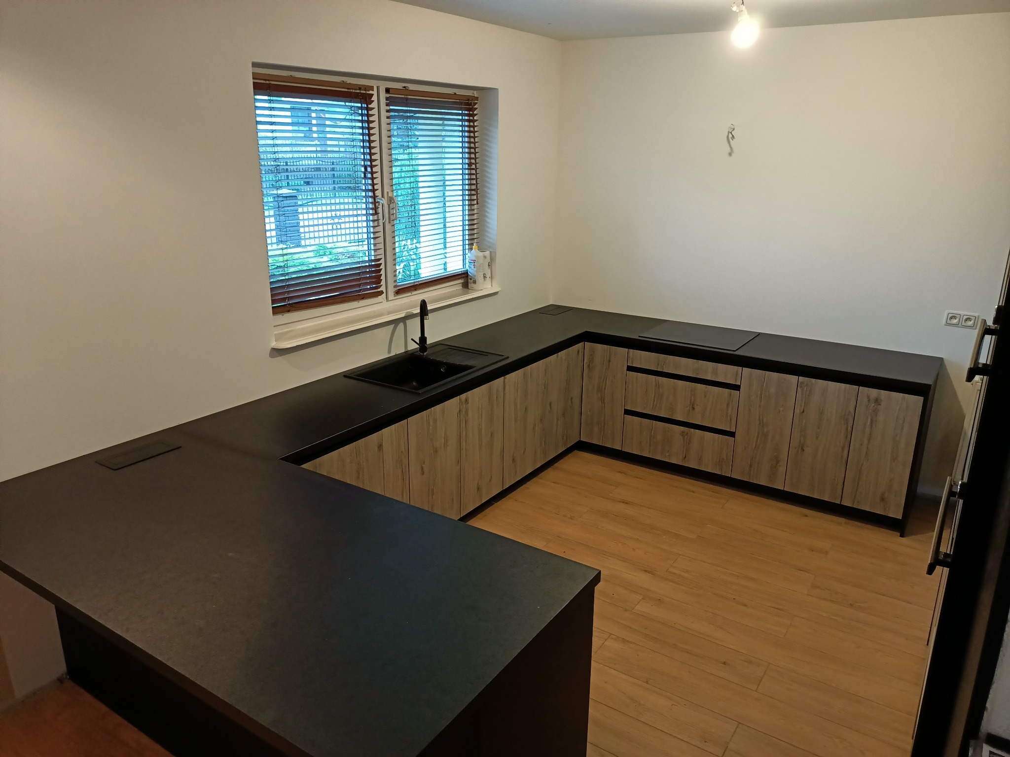 Nowoczesna kuchnia w kształcie litery U z czarnym blatem i drewnopodobnymi frontami szafek, widok na okno z drewnianymi żaluzjami i fragment ściany z wystającymi kablami.