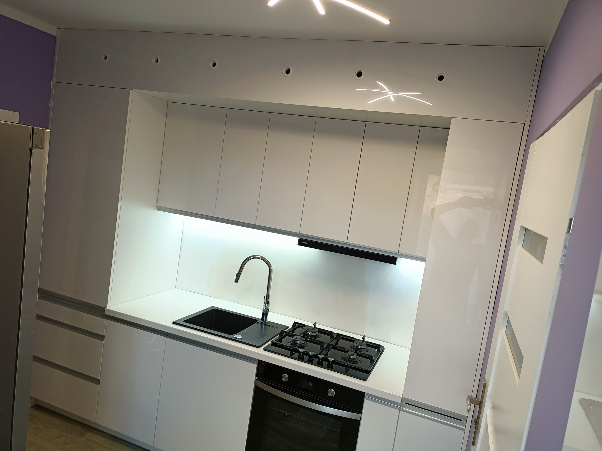 Biała, nowoczesna zabudowa kuchenna z czarnym zlewem i płytą gazową, oświetlenie LED pod szafkami, widoczne elementy wykończenia w kolorze liliowym.