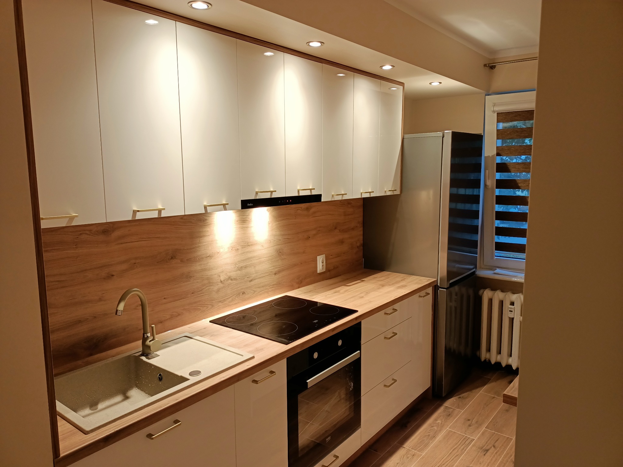 Nowoczesna kuchnia z białymi szafkami, drewnianym blatem i panelem ściennym, z widocznym zlewem granitowym, płytą indukcyjną i piekarnikiem w zabudowie, oświetleniem punktowym oraz lodówką...