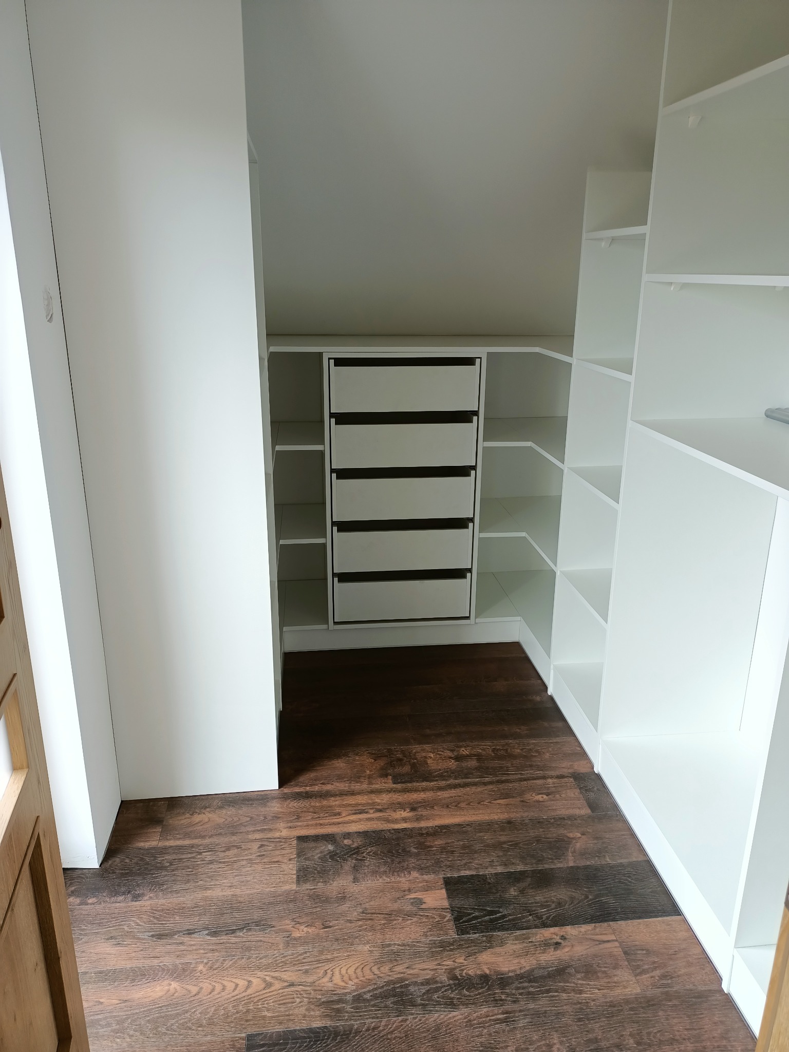 Wnętrze garderoby na poddaszu z białymi półkami i komodą z szufladami z ciemnymi uchwytami, widok z perspektywy wejścia na brązową podłogę.