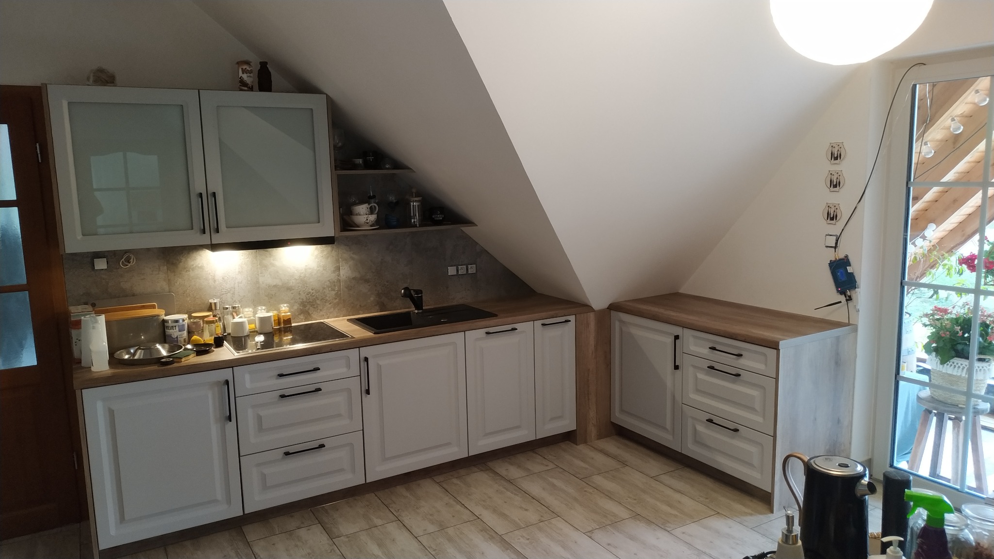 Kuchnia w skosach z białymi frontami szafek, drewnianym blatem i czarnymi uchwytami, z widocznymi półkami i akcesoriami kuchennymi, oświetlona lampą sufitową.