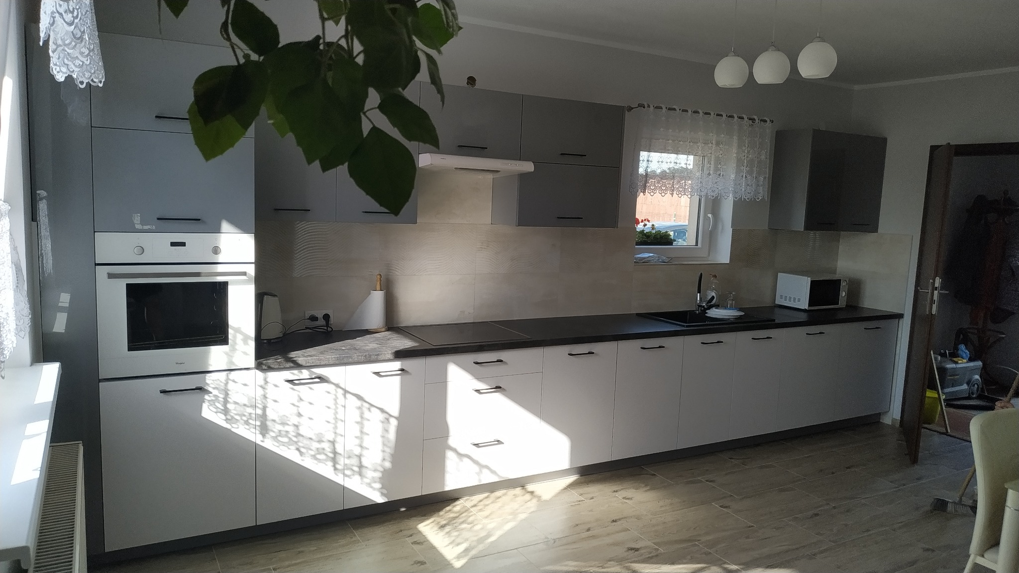 Nowoczesna kuchnia z szarymi i białymi frontami szafek, czarnym blatem, białym piekarnikiem i płytą indukcyjną, oświetlona naturalnym światłem padającym przez okno, z rośliną w tle.