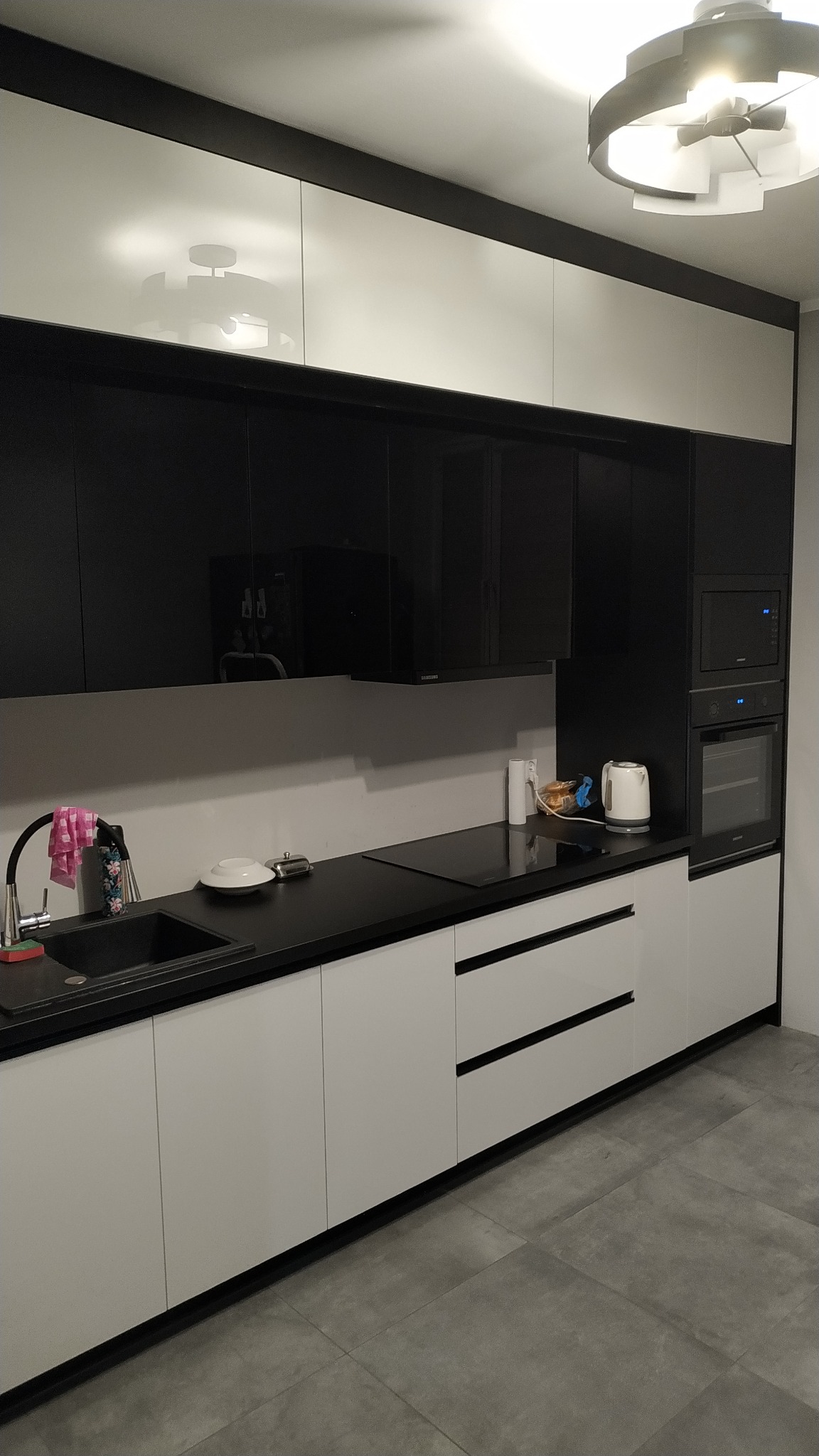 Nowoczesna kuchnia z czarnymi szafkami górnymi na wysoki połysk i białymi szafkami dolnymi z czarnymi uchwytami, czarny blat, płyta indukcyjna i piekarnik w zabudowie, widoczny okrągły plafon.