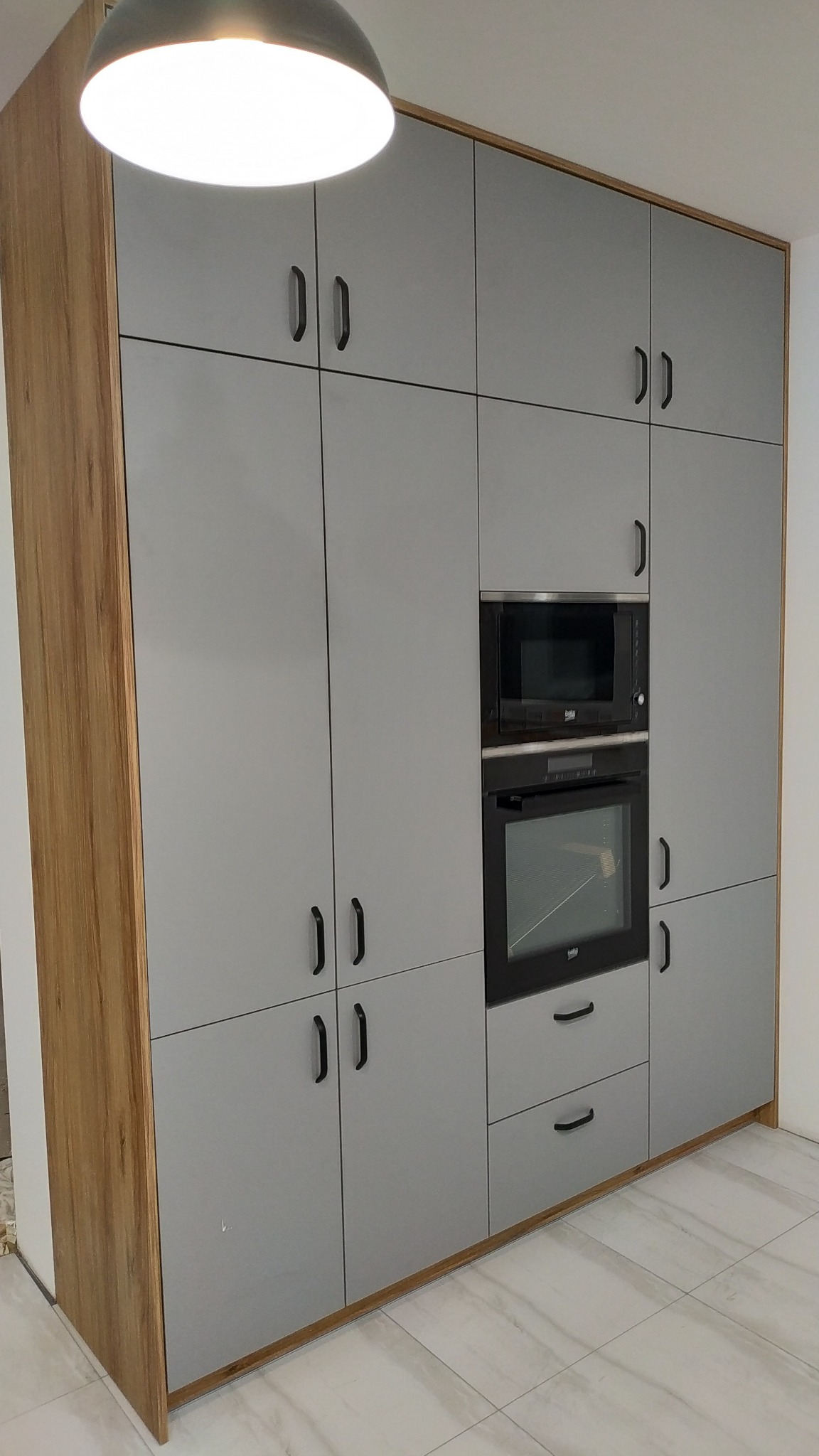 Szara zabudowa kuchenna z drewnianymi bokami, wbudowanym piekarnikiem i mikrofalówką Beko, czarne podłużne uchwyty.