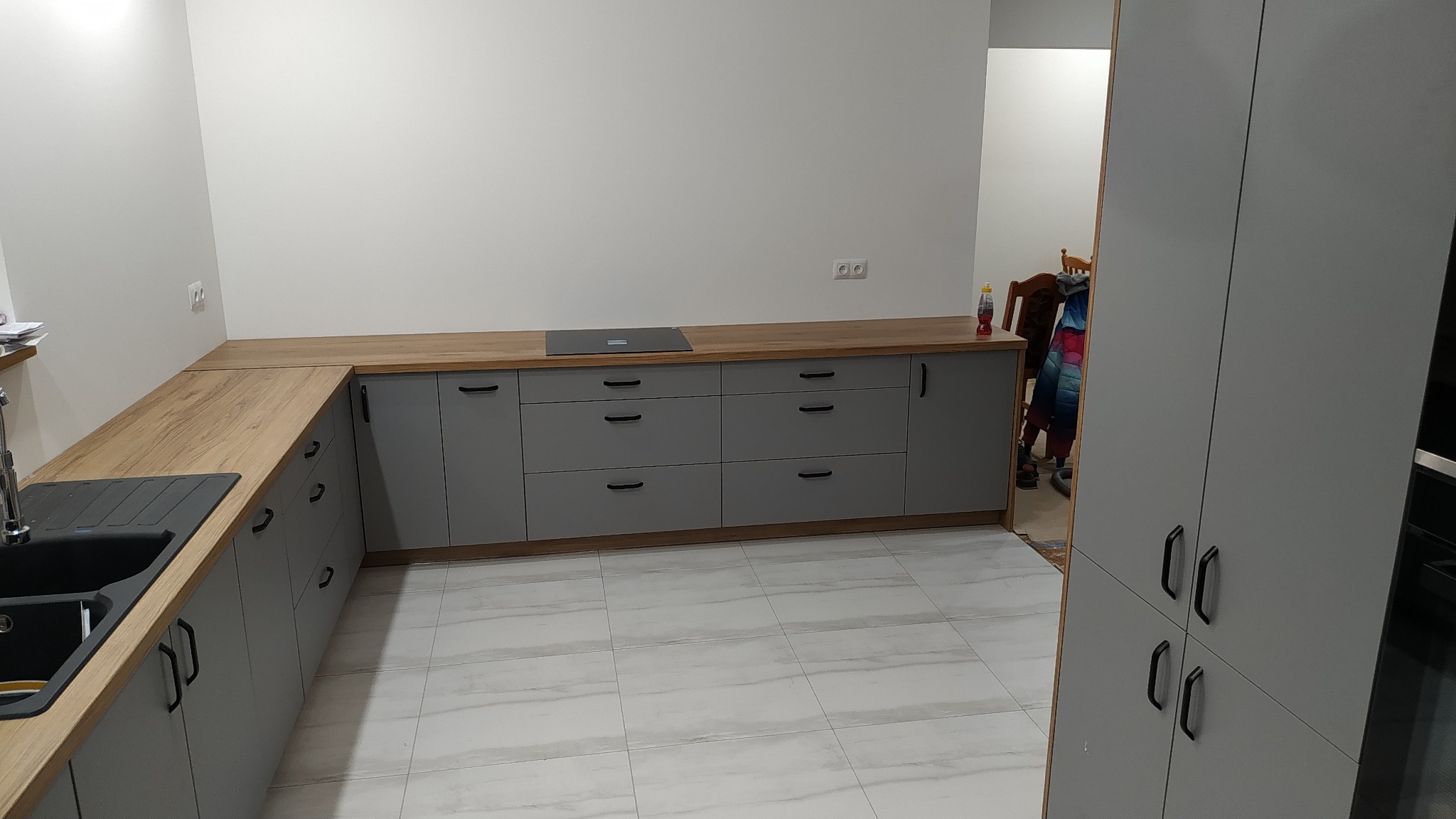 Szara zabudowa kuchenna z drewnianym blatem i czarnym zlewem na tle białych ścian i podłogi z płytek imitujących marmur, widoczna płyta indukcyjna i fragment szafy.