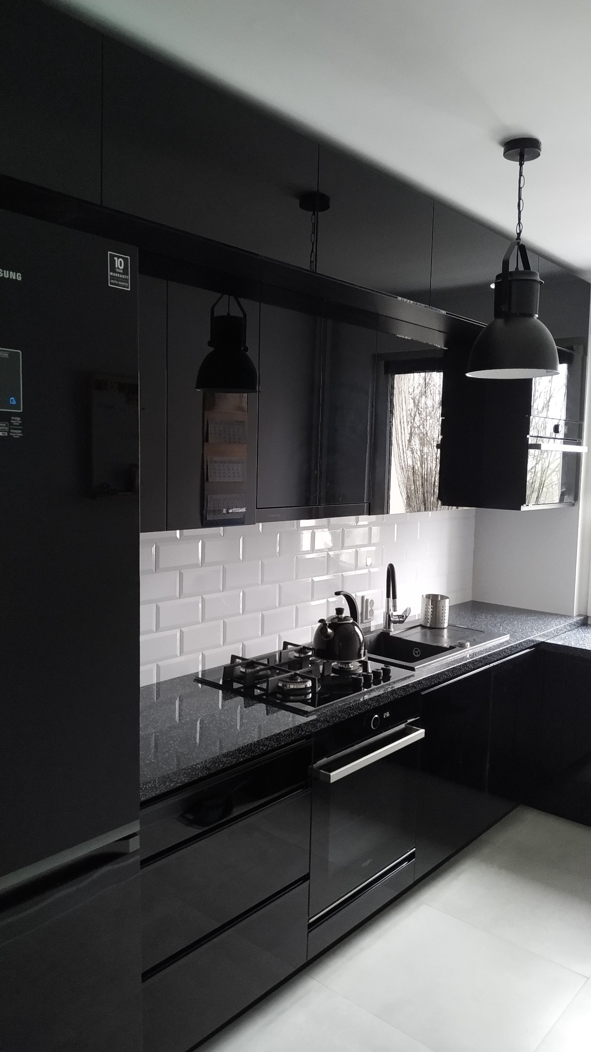 Czarna, błyszcząca zabudowa kuchenna z blatem w czarną mozaikę, białe kafelki na ścianie, czajnik na gazowej płycie grzewczej, wiszące lampy w stylu industrialnym.