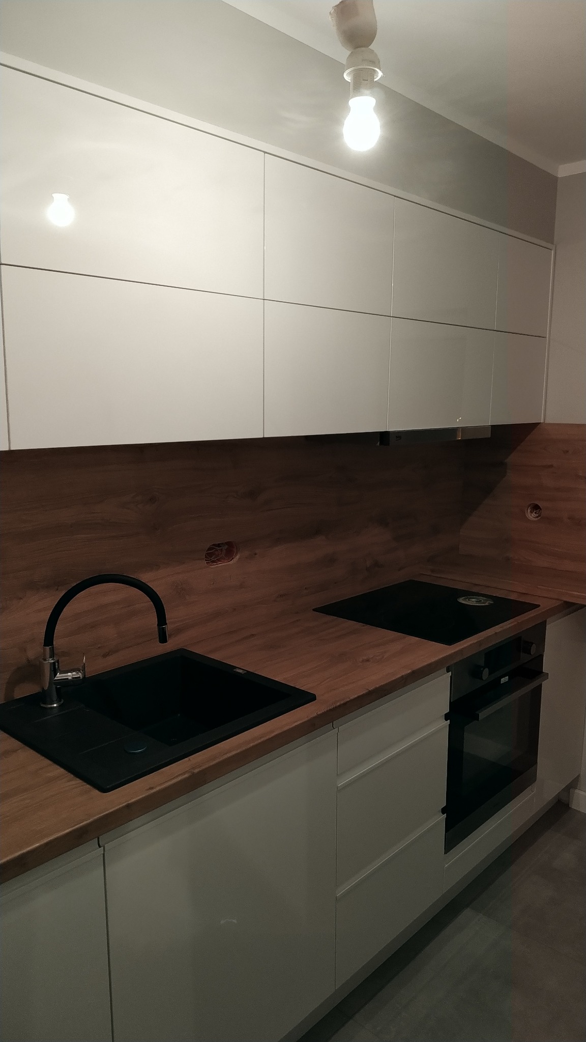 Nowoczesna zabudowa kuchenna w Gorzowie Wielkopolskim z białymi szafkami górnymi i dolnymi, drewnianym blatem oraz czarnym zlewem i płytą indukcyjną. Widoczne niedokończone otwory na gniazdka...