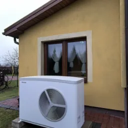 Biała jednostka zewnętrzna pompy ciepła Stiebel Eltron na betonowym fundamencie, ustawiona przed żółtą ścianą domu z brązowym oknem i białymi firankami.