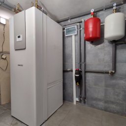 VIAPROFIT SP. Z O.O. SPÓŁKA Z OGRANICZONĄ ODPOWIEDZIALNOŚCIĄ - Biała pompa ciepła marki Stiebel Eltron zainstalowana w pomieszczeniu technicznym z szarymi płytkami, widoczne rury, zbiorniki i skrzynka elektryczna.