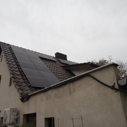 VIAPROFIT SP. Z O.O. SPÓŁKA Z OGRANICZONĄ ODPOWIEDZIALNOŚCIĄ - Dom jednorodzinny z brązową dachówką i panelami fotowoltaicznymi na dachu, widoczny klimatyzator na ścianie, pochmurne niebo.