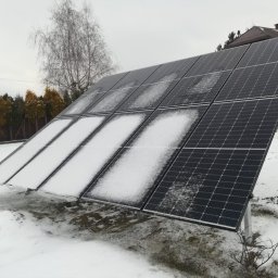 VIAPROFIT SP. Z O.O. SPÓŁKA Z OGRANICZONĄ ODPOWIEDZIALNOŚCIĄ - Panel fotowoltaiczny częściowo pokryty śniegiem na tle ośnieżonego trawnika i żółtego domu w zimowy dzień.