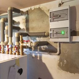 VIAPROFIT SP. Z O.O. SPÓŁKA Z OGRANICZONĄ ODPOWIEDZIALNOŚCIĄ - Węzeł cieplny z licznikiem zużycia energii, rury izolowane termicznie, surowa ściana w tle.