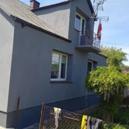 Dom jednorodzinny po remoncie elewacji, pomalowany na szaro, z balkonem z metalową balustradą i anteną telewizyjną na dachu. Na balkonie powiewa flaga Polski. W tle widoczne drzewa i niebieskie...