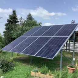 Instalacja fotowoltaiczna na metalowej konstrukcji, widoczna na tle zielonego trawnika, drzew i błękitnego nieba z chmurami.