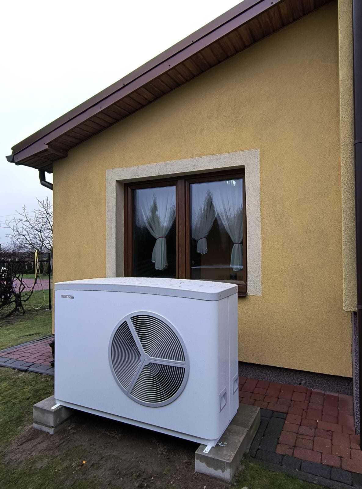 Biała jednostka zewnętrzna pompy ciepła Stiebel Eltron na betonowym fundamencie, ustawiona przed żółtą ścianą domu z brązowym oknem i białymi firankami.