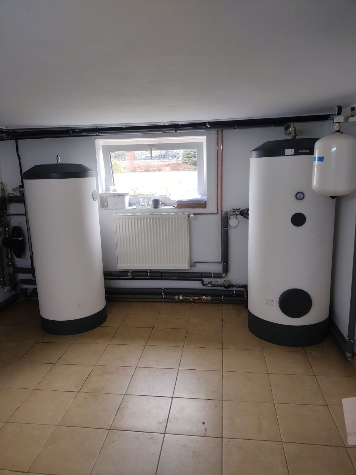 Dwa białe zbiorniki akumulacyjne ciepłej wody w piwnicy z oknem, rurami i grzejnikiem.