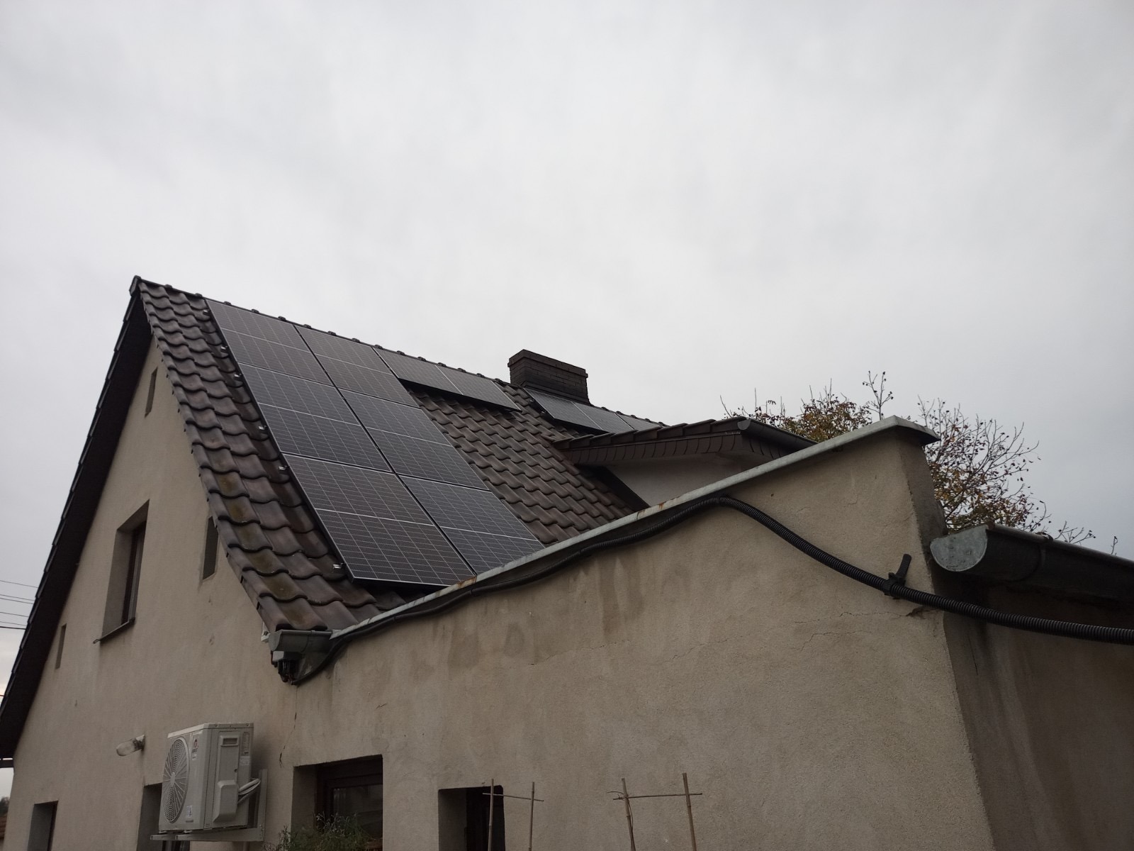 Dom jednorodzinny z brązową dachówką i panelami fotowoltaicznymi na dachu, widoczny klimatyzator na ścianie, pochmurne niebo.