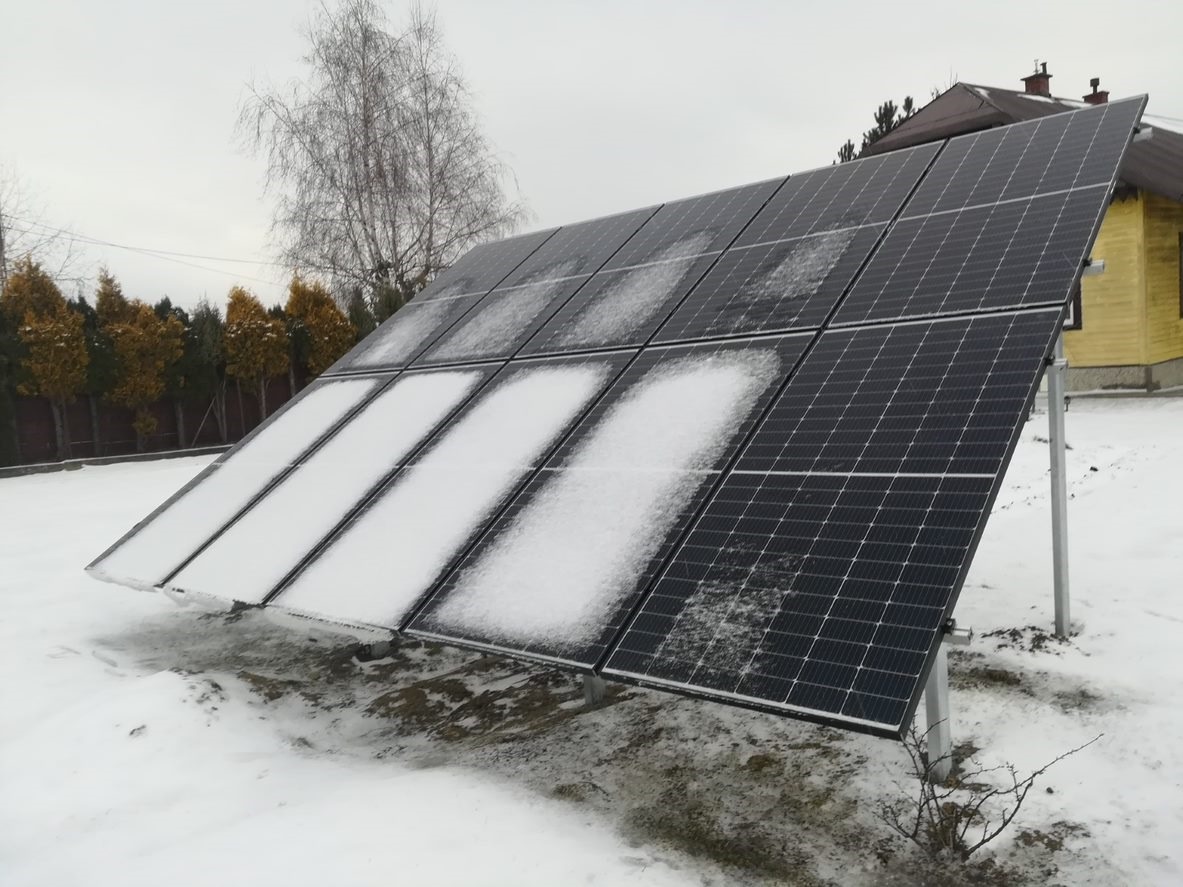 Panel fotowoltaiczny częściowo pokryty śniegiem na tle ośnieżonego trawnika i żółtego domu w zimowy dzień.