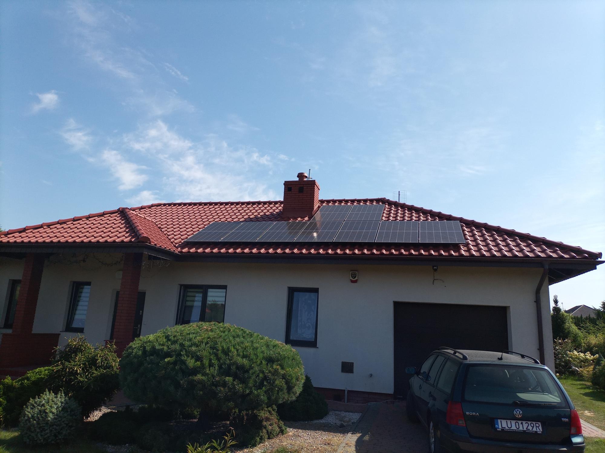 Instalacja fotowoltaiczna na dachu domu jednorodzinnego z czerwoną dachówką, widok z poziomu ogrodu w słoneczny dzień.