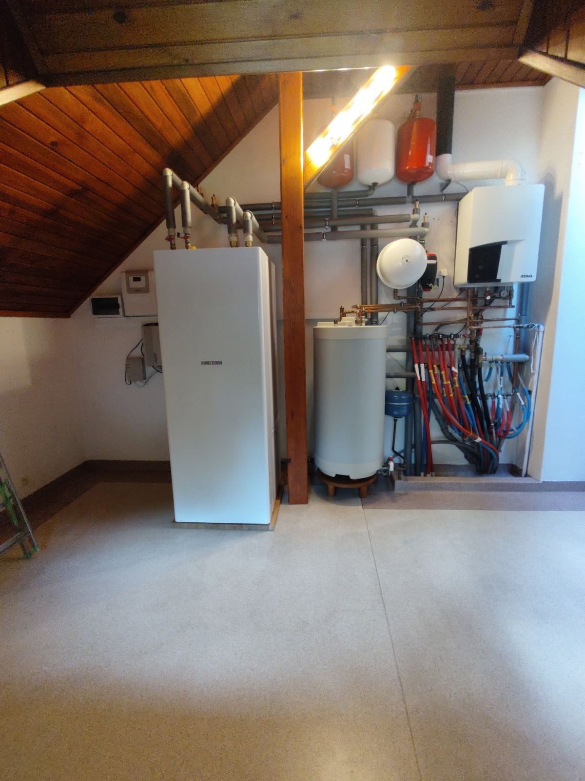 Kompletna instalacja grzewcza w kotłowni pod skośnym dachem, z dwoma kotłami, zasobnikiem ciepłej wody i rozbudowanym systemem rur i przewodów, w tym czerwonych i niebieskich.