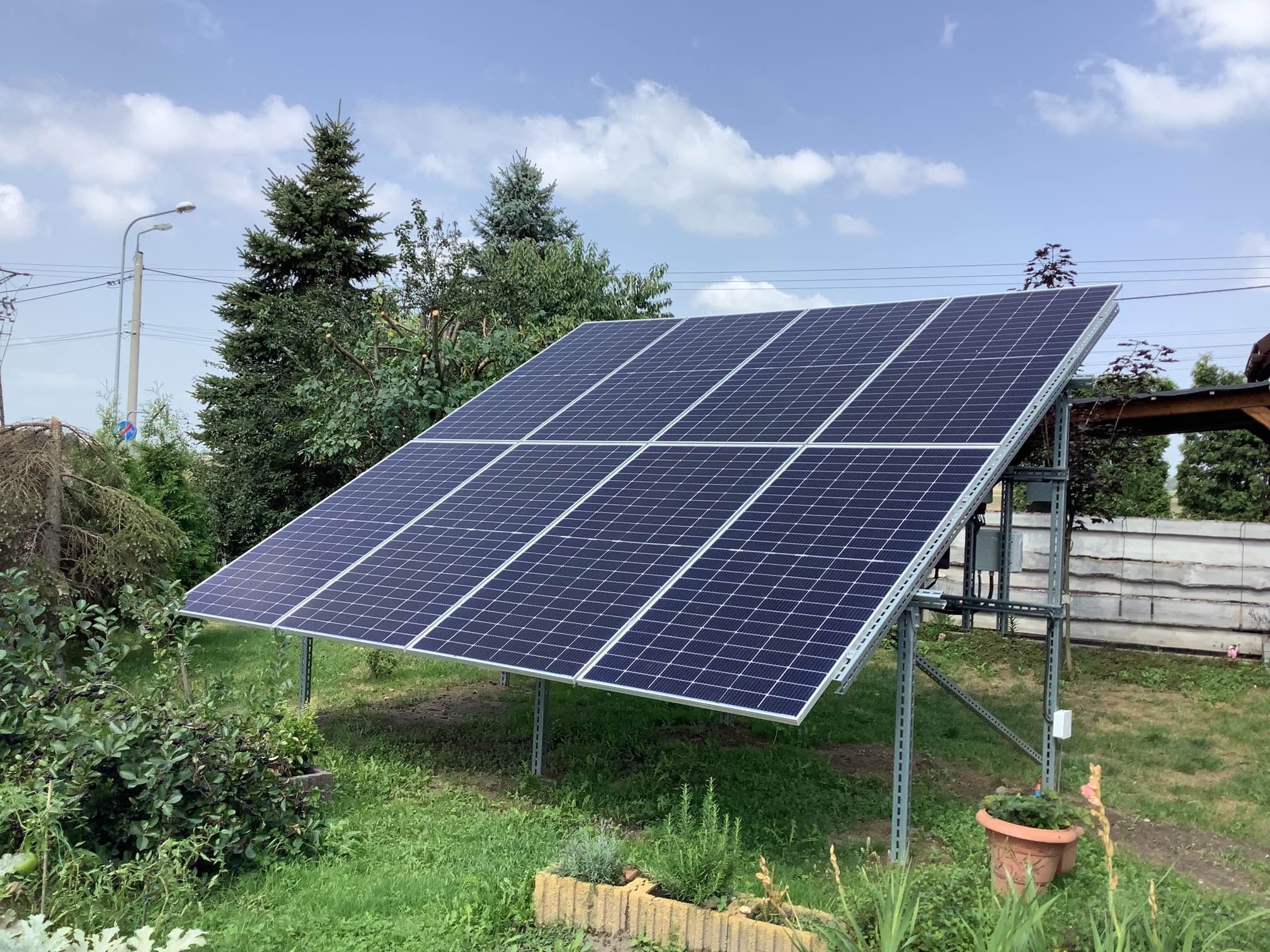Instalacja fotowoltaiczna na metalowej konstrukcji, widoczna na tle zielonego trawnika, drzew i błękitnego nieba z chmurami.