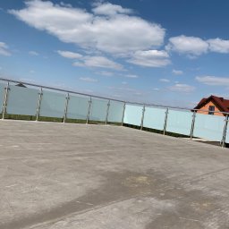 Steel Art Balustrady Nierdzewne - Szklana balustrada z nierdzewnymi słupkami, zamontowana na tarasie w Katowicach, widok z dołu na niebieskie niebo z chmurami i dom w tle.