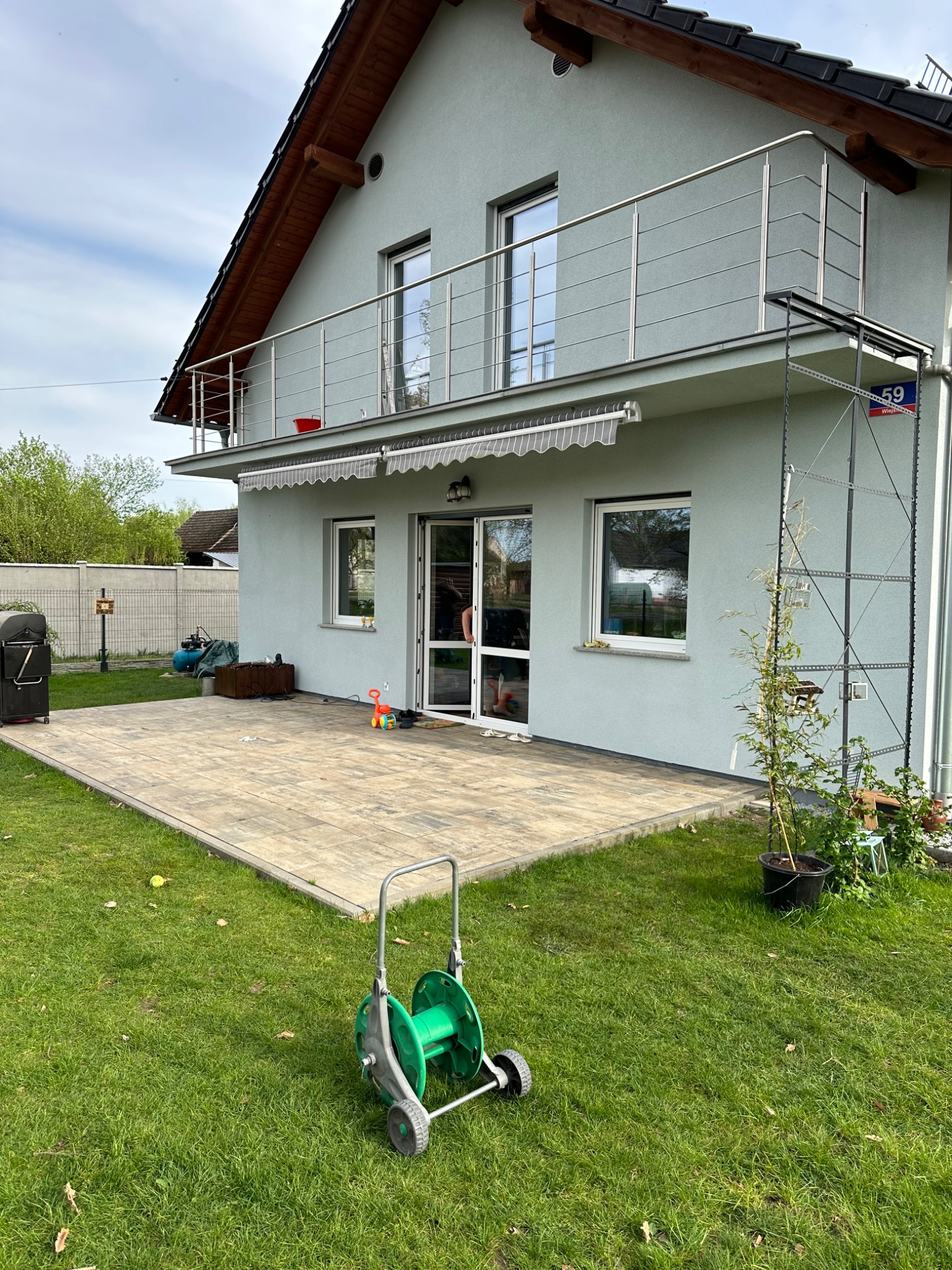 Dwukondygnacyjny dom jednorodzinny z balkonem z nowoczesną metalową balustradą, widok z ogrodu na taras wyłożony kostką brukową.