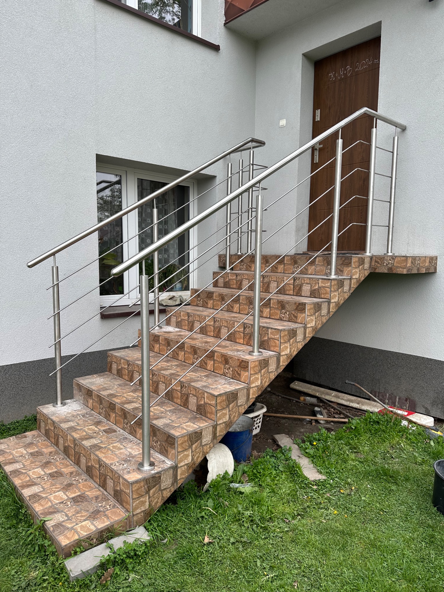 Zewnętrzne schody wejściowe wyłożone brązową mozaiką, z prostą balustradą ze stali nierdzewnej, widoczne narzędzia budowlane i wiadro w tle.