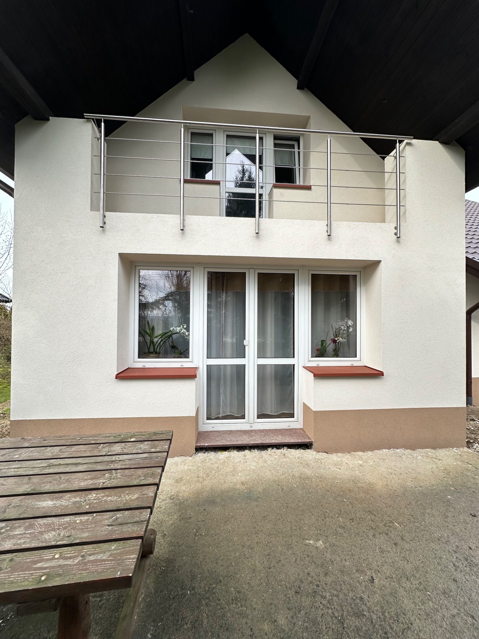 Dwupiętrowy budynek z balkonem zabezpieczonym metalową, poziomą konstrukcją, widoczne okna z kwiatami w doniczkach, drewniany stół na pierwszym planie.