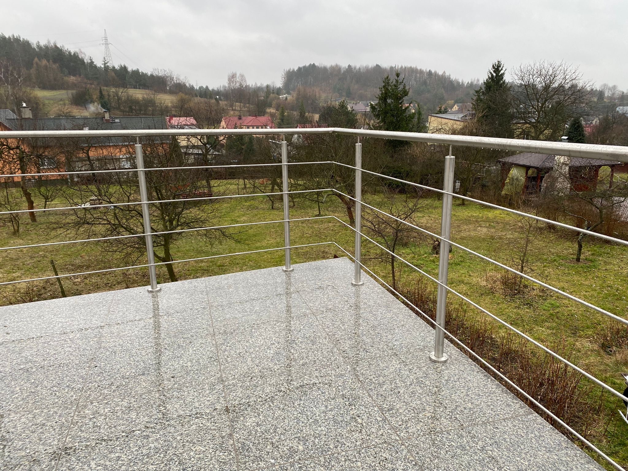Balkon z szarą, granitową posadzką i nowoczesną balustradą ze stali nierdzewnej z widokiem na zalesione wzgórza w pochmurny dzień.
