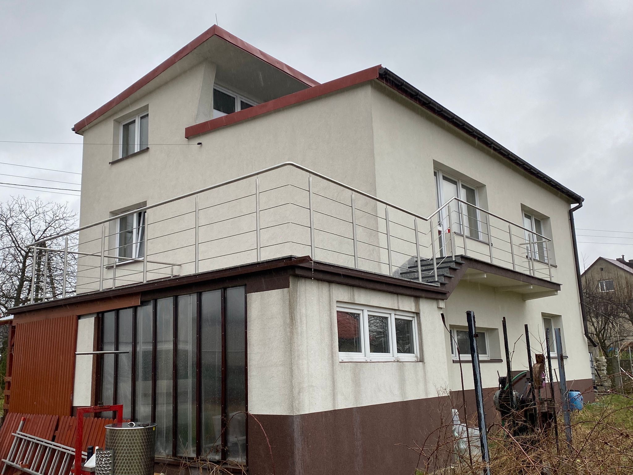 Dwupiętrowy dom z balkonem zabezpieczonym metalową balustradą z poziomych prętów, widoczny z zewnątrz w pochmurny dzień.